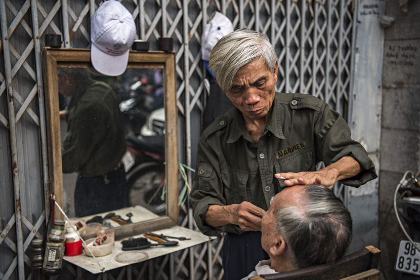 Una delle tante attività svolte sui marciapiedi di Hanoi: un barbiere vestito con una mimetica militare  americana alle prese con un cliente